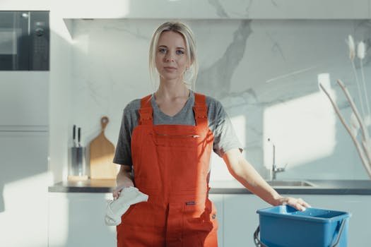 A woman in an orange apron stands in a modern kitchen for her cleaning business.